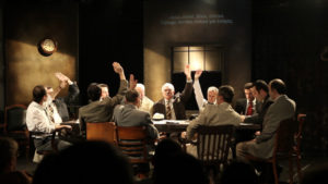 γύρω από ένα μεγάλο ορθογώνιο τραπέζι στο κέντρο μιας θεατρικής σκηνής είναι καθισμένοι 12 άντρες με κοστούμια. Οι 6 από αυτούς έχουν σηκωμένο το χέρι τους. Στο πίσω μέρος της σκηνής πάνω στον τοίχο προβάλλονται υπέρτιτλοι.