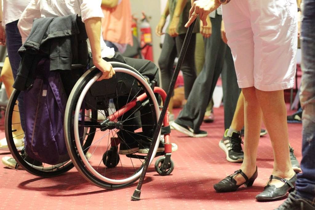A group of people moves into a room with red carpet. Their bodies can be seen from the waist down as they move through the space. Some move with a wheelchair, some with crutches and some without assistance.