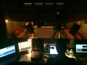 στο ηλεκτρολογείο ενός θεάτρου, η πλάτη μιας γυναίκας στο σκοτάδι μπροστά από ένα μικρόφωνο. Μπροστά της τέσσερις ανοιχτές οθόνες και πίσω από το τζάμι μπροστά της η άδεια αίθουσα ενός μεγάλου θεάτρου με κόκκινα καθίσματα