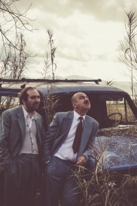 δύο άντρες με παλιά κοστούμια δίπλα από ένα εγκαταλειμένο αυτοκίνητο