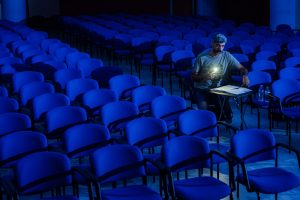 ανάμεσα στα μπλε καθίσματα μιας σκοτεινής θεατρικής αίθουσας ένας άντρας με φακό φωτίζει ένα κείμενο