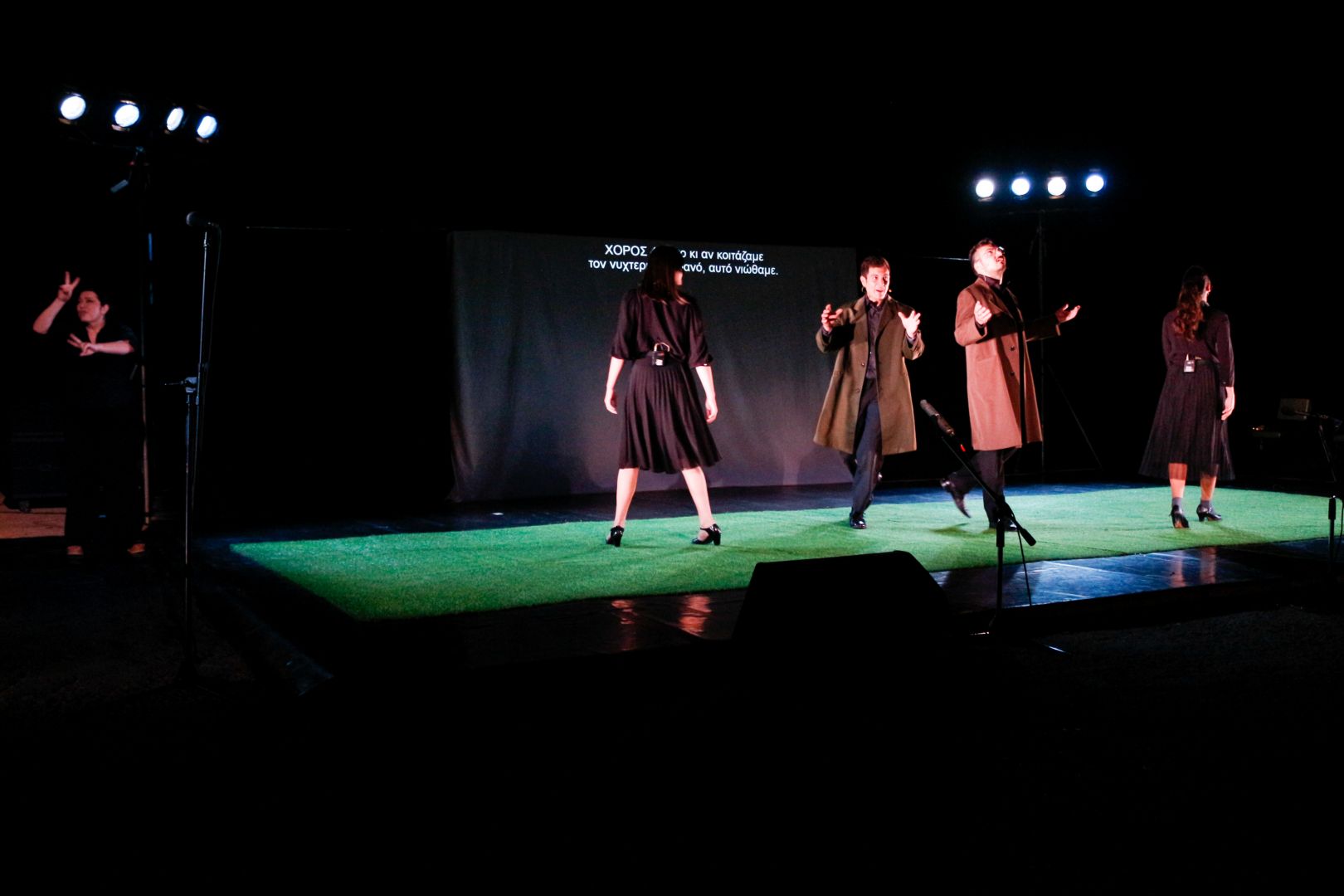 σε μία θεατρική σκηνή με πράσινο πάτωμα τρία άτομα βηματίζουν με μεγάλους δρασκελισμούς ενώ στο πίσω μέρος προβάλλονται υπέρτιτλοι