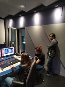 σε ένα στούντιο ηχογράφησης δύο γυναίκες και ένας άντρας κοιτούν προς μία ανοιχτή οθόνη όπου προβάλλεται ένα βίντεο, μπροστά από την οθόνη η μία γυναίκα χειρίζεται μία κονσόλα ήχου