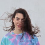 μία κοπέλα με χρωματιστή μπλούζα σε παστέλ αποχρώσεις χαμογελάει ελαφρά καθώς ο αέρας της ανακατεύει τα καστανά της μαλλιά