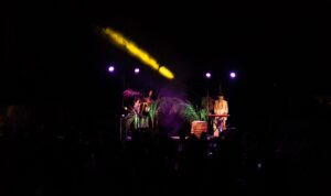Ένας κιθαρίστας με ανοιγμένο πουκάμισο και τζιν παντελόνι και ένας πιανίστας με καλυμμένο το πρόσωπο με ένα χρυσαφί βέλο παίζουν μουσική σε μία σκηνή φωτισμένη με απαλά μωβ και κίτρινα φώτα και στολισμένη με μικρούς φοίνικες.