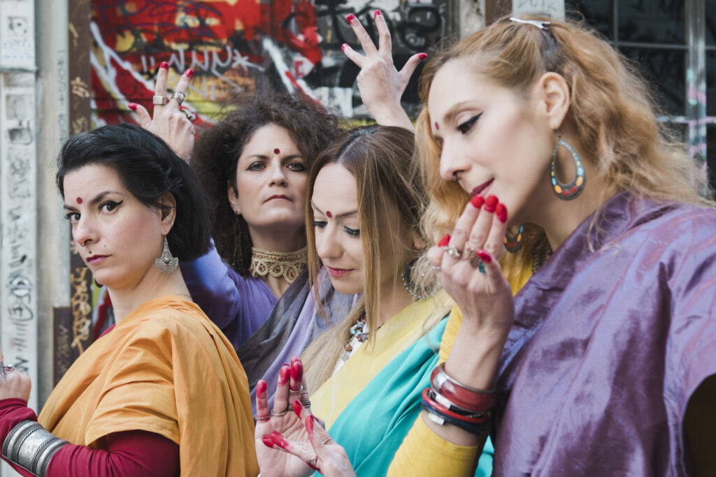Τέσσερις νέες γυναίκες με πολύχρωμα ινδικά σάρι, κοσμήματα και κόκκινη τιλάκα στα μέτωπά τους στέκονται η μία κοντά στην άλλη σε ένα αστικό τοπίο με γκραφίτι. Οι δύο κοιτούν με ένταση το φακό ενώ οι άλλες δύο έχουν χαμηλωμένο το βλέμμα και μοιάζουν σκεπτικές. Τα ακροδάχτυλα όλων είναι βαμμένα κόκκινα. 