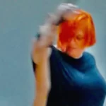 Μία γυναίκα με έντονα κόκκινα μαλλιά σε καρέ και γυαλιά οράσεως έχει το δεξί της χέρι σηκωμένο ψηλά και στρέφει το πρόσωπο χαμηλά καθώς μοιάζει να χορεύει.