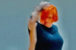 Μία γυναίκα με έντονα κόκκινα μαλλιά σε καρέ και γυαλιά οράσεως έχει το δεξί της χέρι σηκωμένο ψηλά και στρέφει το πρόσωπο χαμηλά καθώς μοιάζει να χορεύει.