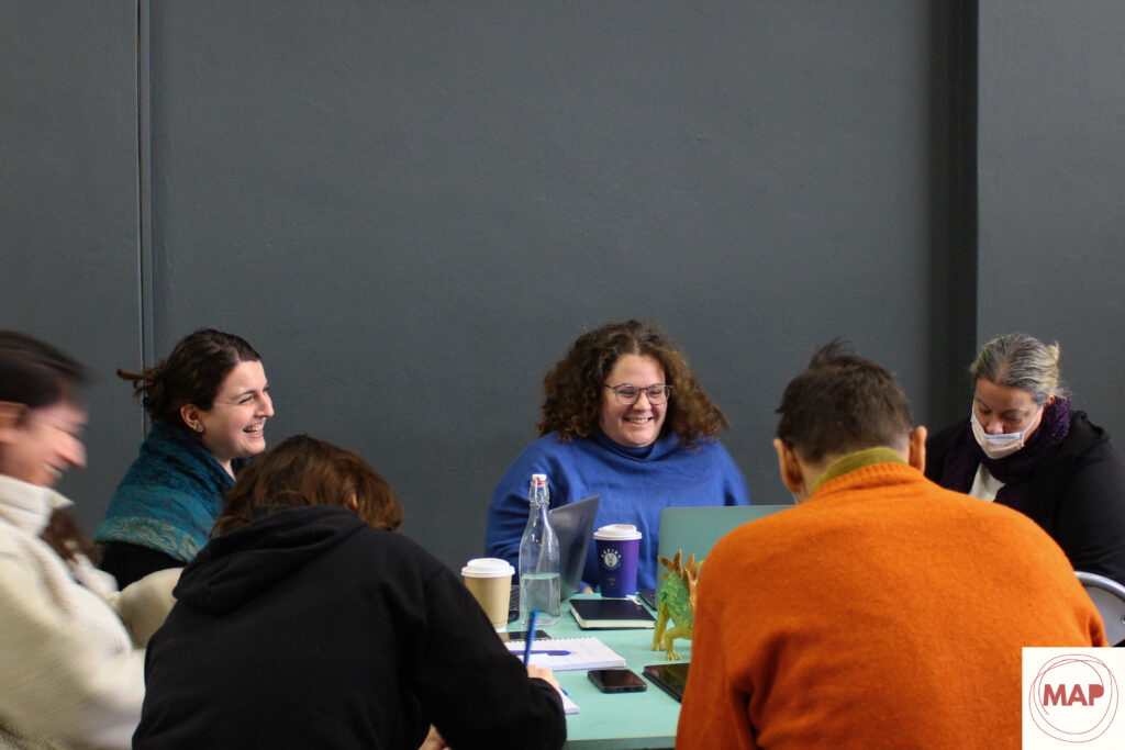 Μια ομάδα ανθρώπων είναι καθισμένη γύρω από ένα τραπέζι σε έναν χώρο με γκρι τοίχους. Γελούν καθώς