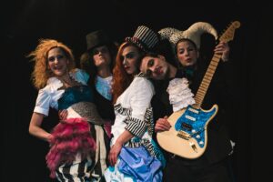 Σε μαύρο φόντο, τέσσερις γυναίκες κι ένας άντρας με ηλεκτρική κιθάρα ποζάρουν όρθιοι. Φορούν κοστούμια με φραμπαλάδες, σακάκια, κορσέδες και φούστες με πιέτες και στα κεφάλια τους μεγάλα καπέλα με παράξενα σχήματα.