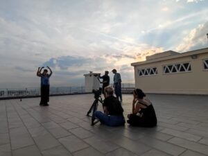 Σε μια μεγάλη ταράτσα στο κέντρο της Αθήνας, ο ήλιος δύει στο βάθος. Στα αριστερά, ένα ζευγάρι χορευτών εκτελεί μια χορογραφία σε ντουέτο ενώ γύρω τους κάμερες και άτομα του συνεργείου τους βιντεοσκοπούν. 