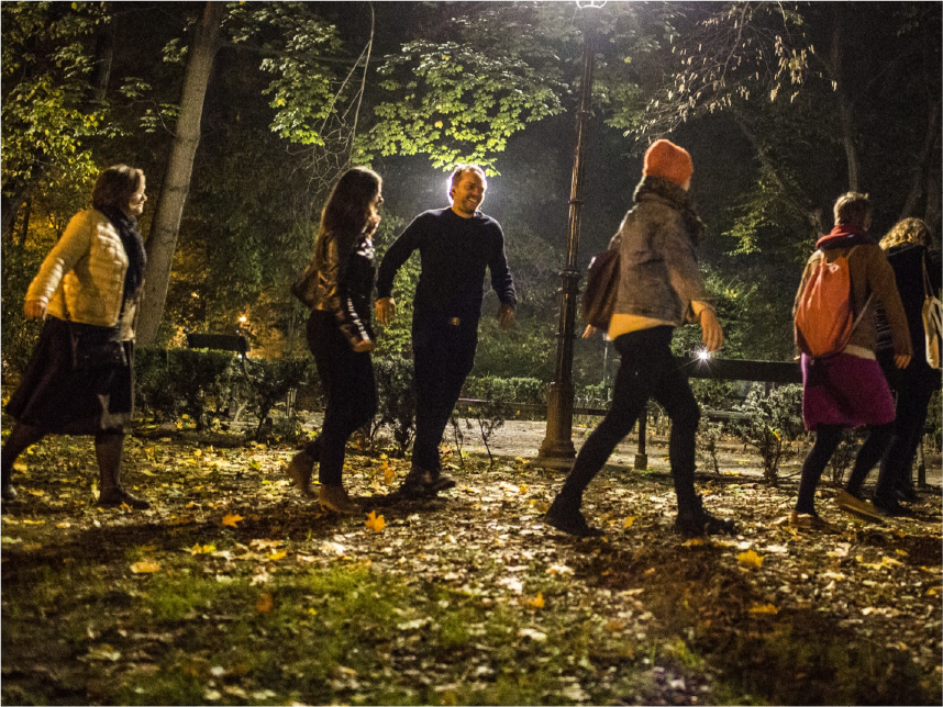 Σε νυχτερινό τοπίο σε πάρκο, ολόσωμη φωτογραφία μιας ομάδας έξι νέων σε προφίλ με σακίδια στις πλάτες και χειμερινά ρούχα που περπατά. Ένας από αυτούς είναι ελαφρά στραμμένος προς την κάμερα και φαίνεται η χαρούμενη έκφραση στο πρόσωπό του. 