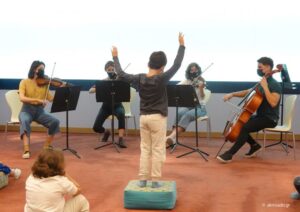 Σε έναν χώρο με μοκέτα ένα παιδί γύρω στα 10 στέκεται όρθιο πάνω σε ένα μαξιλάρι με τα χέρια ψηλά σε κίνηση σα να διευθύνει μια ορχήστρα. Μπροστά του τέσσερις μουσικοί με έγχορδα όργανα τον κοιτούν και παίζουν.