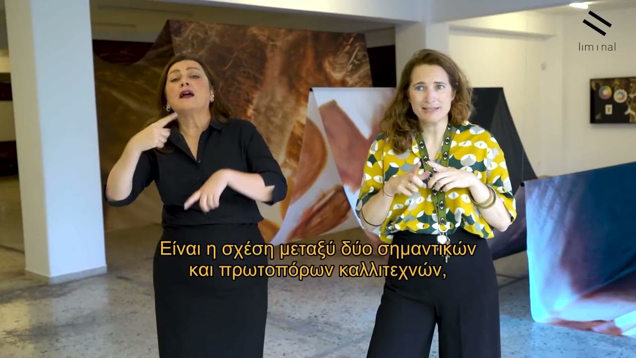 In an exhibition space, a tour guide dressed in a yellowish shirt and black trousers presents the content of the exhibition. Beside her, the sign language interpreter dressed in black is simultaneously translating into sign language. The photo is an excerpt from a video, at the bottom of which there is a subtitle for deaf and hard of hearing people where the words of the tour guide are rendered. It is written: "It is the relationship between two important and pioneering artists." Contact us