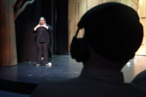 Φωτογραφία ενός άνδρα από πίσω ενόσω παρακολουθεί μία παράσταση φορώντας ακουστικά. Στη σκηνή μπροστά του μια διερμηνέας της νοηματικής αποδίδει το περιεχόμενο της παράστασης για κωφά και βαρήκοα άτομα.