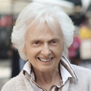 Πορτραίτο μιας γυναίκας γύρω στα 70 με λευκά μαλλιά και μεγάλα καστανά μάτια που χαμογελά κεφάτα στον φακό.
