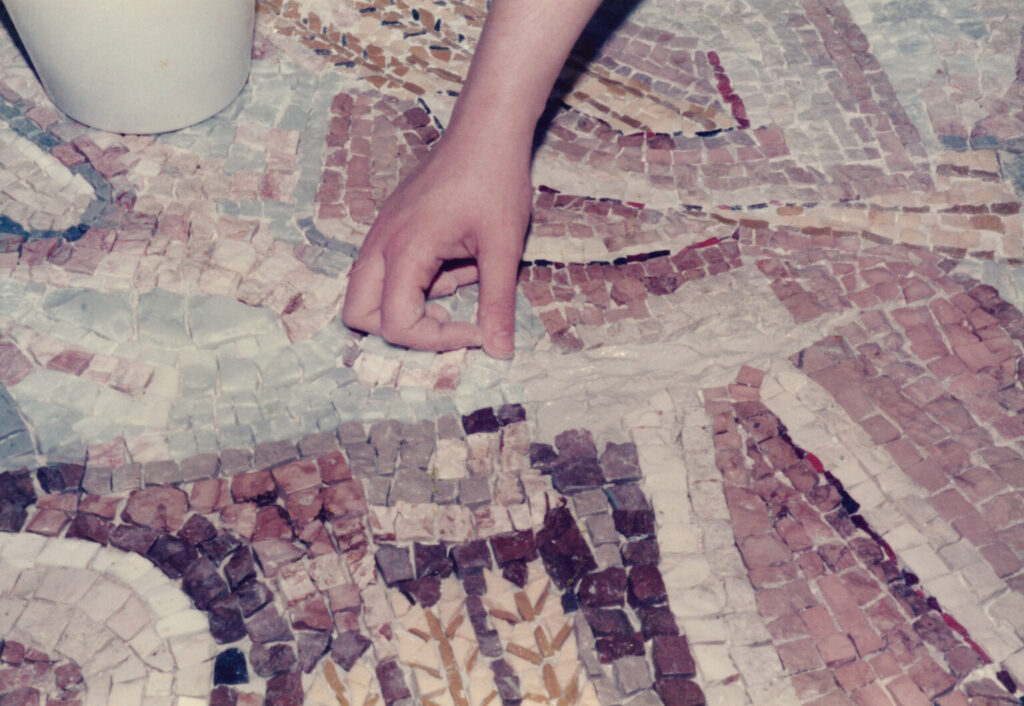 In an old faded color photograph, a hand places a tile in a large mosaic work on the floor.