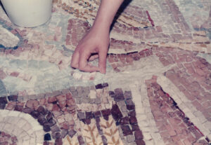Σε μια παλιά έγχρωμη αλλά ξεθωριασμένη φωτογραφία, ένα χέρι τοποθετεί μία ψηφίδα σε ένα μεγάλο ψηφιδωτό έργο στο πάτωμα.