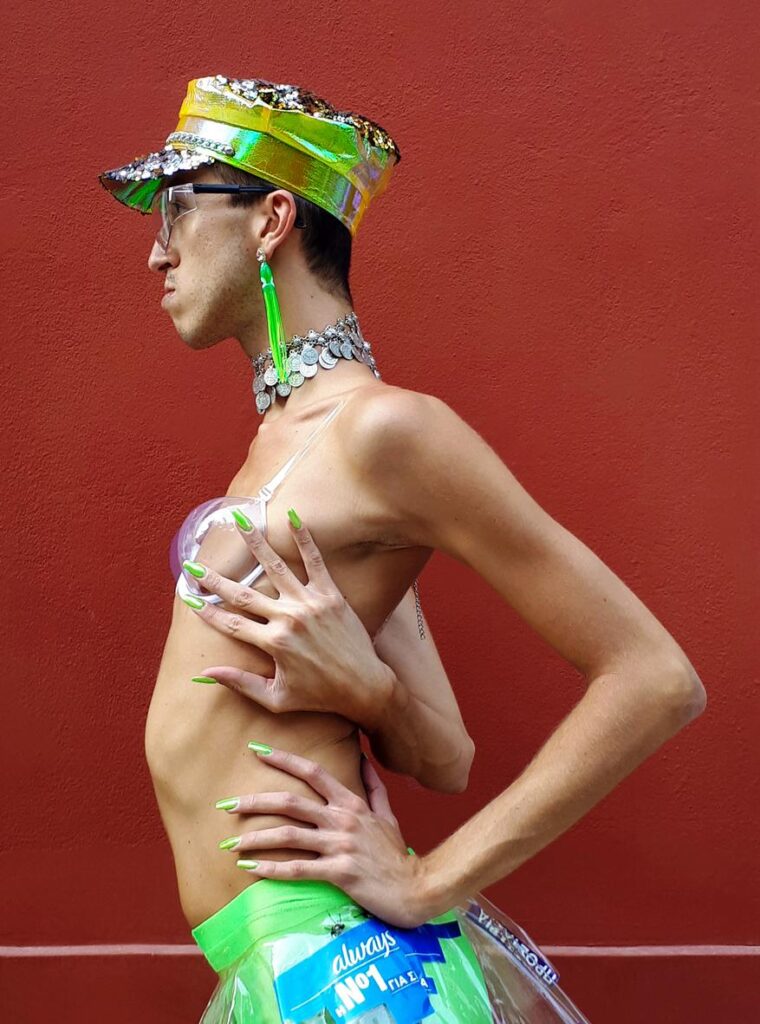In front of a deep red wall poses in profile a young queer-looking man. He wears a soft jockey hat with rhinestone rhinestones, transparent large glasses, a long lime green earring and a silver necklace with rows of round sequins. His naked skinny torso is covered only by a transparent bra with a round hard cup on the chest and a pair of lime green leggings lined with plastic and nylon pieces. His impressively long delicate arms with painted lime green nails weave around his torso and waist forcing his body into a graceful backward bend. 