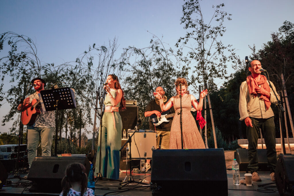 In an open space at dusk, three men and two women dressed in summer clothes in pastel shades share a musical stage. Some sing in front of microphones, others play musical instruments like guitar and bass, and everyone looks cheerful and smiling.