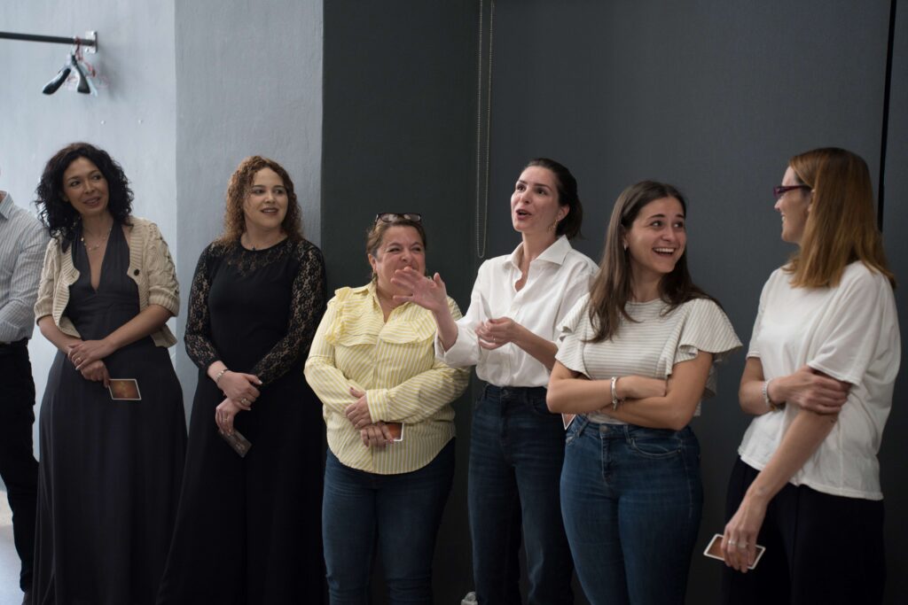 Six women are standing in a room with grey walls. One of them, in a white shirt and brown hair in a ponytail, speaks with a smile and waves her hands as if explaining something. Next to her, a shorter woman in a yellow shirt looks at her and laughs. On the far left, two women in long dresses watch her carefully, while on the left side of the photo, the two women next to her share a comment and laugh as if in agreement.