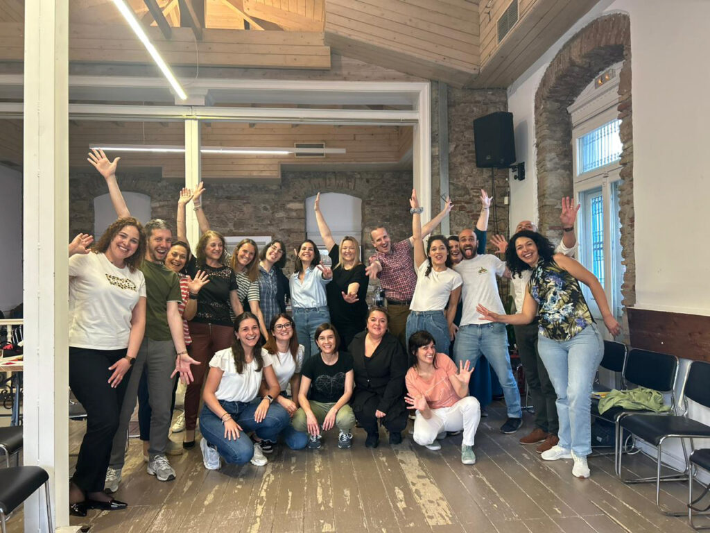 Group photography in a high-ceilinged room with a wooden floor and stone walls with large windows. The group members, mostly women, in summer clothes and smiling faces, raise their hands in a broad salute.
