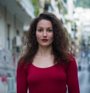 Μια νεαρή κοπέλα με καστανά σπαστά μαλλιά ως το στήθος φορά μπορντό μακρυμάνικη μπλούζα που ταιριάζει με το κραγιόν της. Έχει λεπτά χαρακτηριστικά, μεγάλα καστανά αμυγδαλωτά μάτια και λευκό δέρμα. Πίσω της θολό το αστικό τοπίο.