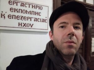 Ένας νέος άντρας με τραγιάσκα και κασκόλ στέκεται μπροστά από μια επιγραφή που γράφει με εκκλησιαστικούς χαρακτήρες Εργαστήριο Εκπομπής και Επεξεργασίας Ήχου.