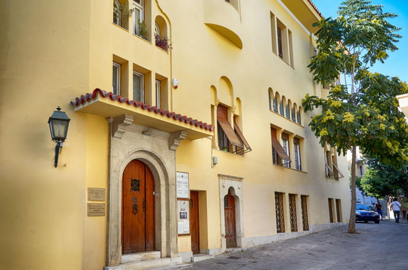 Exterior view of the Museum "Angeliki Chatzimichali" in a paved street of Plaka. The building, created by the architect Aristotle Zachos, is a two-storey building in yellowish colour and is distinguished by its special architecture that combines traditional elements found in Macedonian architecture and in the neo-Byzantine tradition, such as the wooden door frames, the carved frames around the windows or doors and the tiled roof.