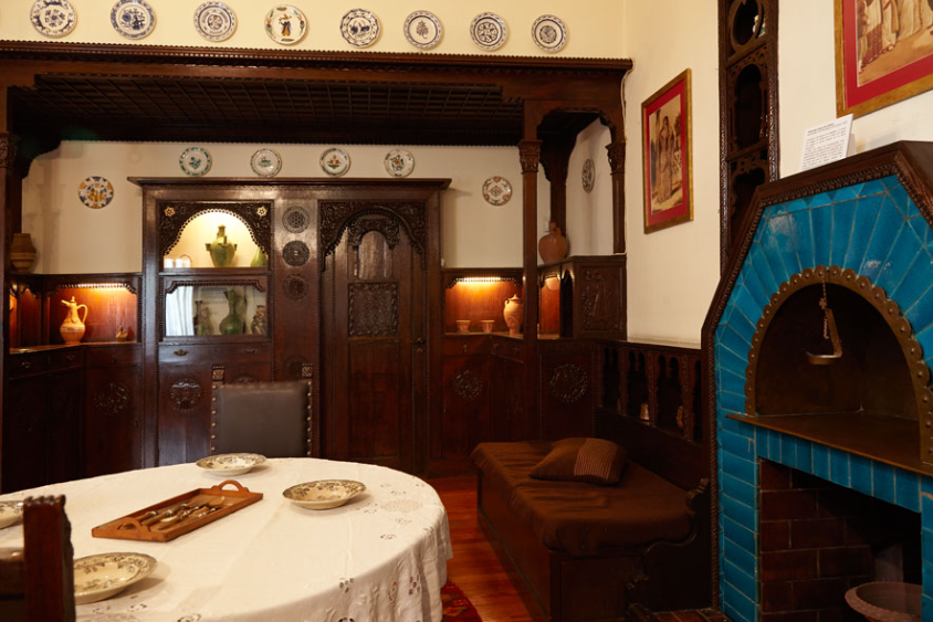 A room inside the Chatzimihali Museum that resembles a salon of the early 19th century. In the centre there is a table covered with a white tablecloth, on the left a chest that also serves as a divan and next to it a fireplace with blue tiles. In the background, around a large wooden piece of furniture hanging on the walls like a display case, are a number of colourful traditional plates.