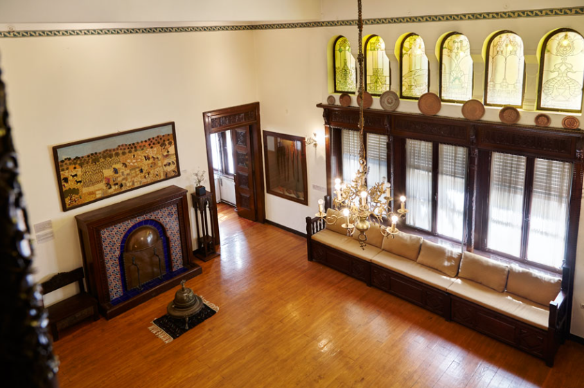 Panoramic view of a room with a wooden floor inside the Chatzimihali Museum. At the spacious entrance of the museum, we find an old fireplace decorated with coloured tiles and a large sofa consisting of wooden carved divan chairs with beige cushions. Plenty of light comes in through the tall windows at the bottom and smaller windows adorned with stained glass near the high ceiling.