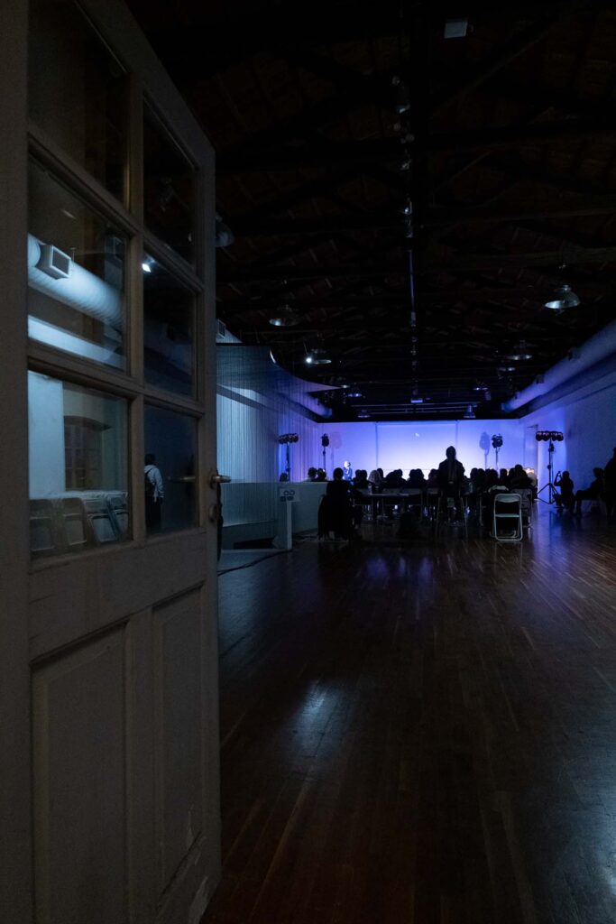 An open white glazed door leads to a large long room with a wooden floor. In the background you can vaguely see a crowd of people sitting in chairs as if watching something under a soft purple lighting.