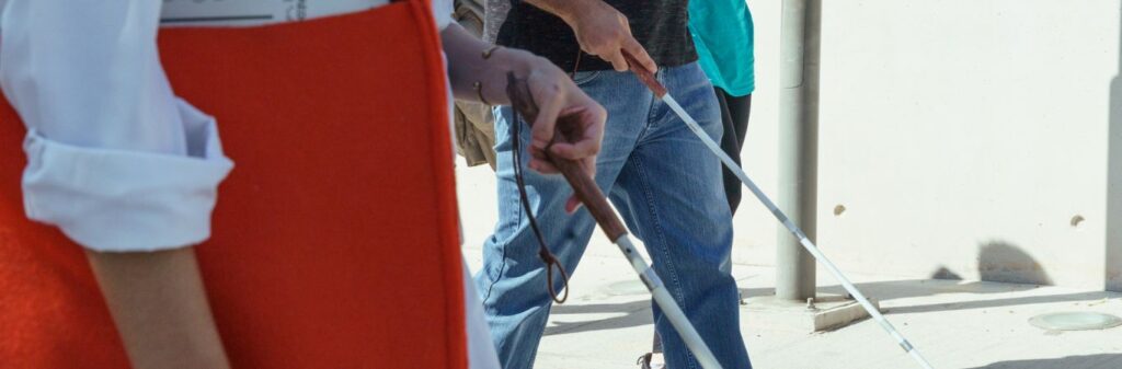 White cane users move in a sunny outdoor space with a grey wall. The photo focuses on the height of the hands holding the cane, reaching to about their knees.