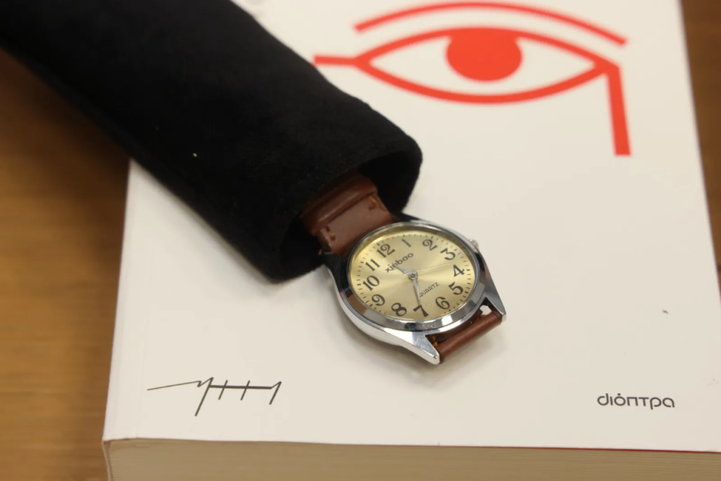 On the cover of a book by Kazantzakis published by Dioptra is a thin black cloth case. From within it emerges an analogue wristwatch with a brown leather strap.