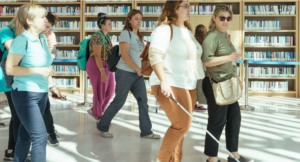 Σε μία βιβλιοθήκη με μαρμάρινο πάτωμα που λούζεται από το πρωινό φως, μια ομάδα γυναικών με ανοιξιάτικα ρούχα προχωρά κατά μήκος των ραφιών με τα βιβλία στο βάθος. Ανάμεσά τους βρίσκεται και μια νεαρή γυναίκα που προχωρά με το λευκό της μπαστούνι κρατώντας από το μπράτσο μια άλλη γυναίκα στα αριστερά της.