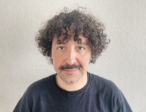 A man with curly brown curls falling on his forehead and a moustache looks at the camera with a soft smile.