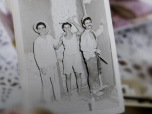 Ένα γερασμένο χέρι κρατά μια ασπρόμαυρη μικρή φωτογραφία. Σε αυτήν, τρεις νεαροί άνδρες με πηλοφόρια ποζάρουν χαμογελαστοί μπροστά από μισοσοβατισμένους τοίχους.