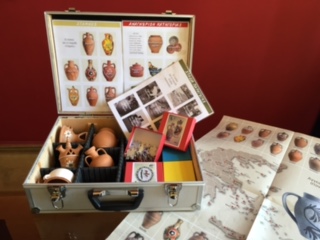 An open suitcase with a hard metal casing, the museum equipment, lies open, revealing small replicas of vessels, brochures, catalogs, and boxes inside.