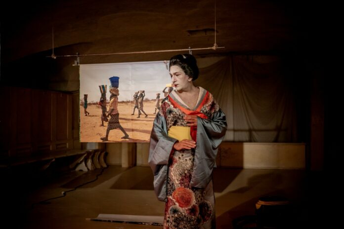 In a room with a light-colored wooden floor a man with male features disguised as a geisha stands upright with a stern look, staring at something down right as if something has caught his attention. His black hair is caught in an elaborate high bun, his skin is dyed white and his lips are red. He wears a shiny kimono with wide gray sleeves and a pattern of blooming chrysanthemums stretching all the way down. Behind him on a projection screen are seen lean bodies of black people in a desert landscape carrying baskets on their heads.