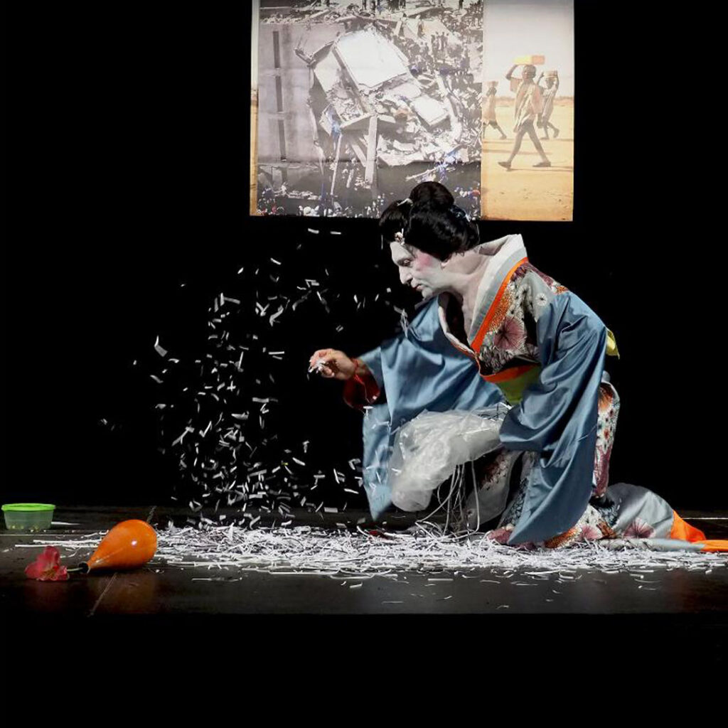 On a stage, a geisha with her face painted white and her black hair caught in an elaborate bun is kneeling in profile over thin strips of white paper covering the black floor. With a wave of her hand that tugs at the wide sleeve of her colorful kimono she lifts the small pieces of paper in front of her creating a paper snowstorm. Behind her in a video projection are vague images of piles of objects as if in a garbage dump and young black boys carrying square baskets on their heads across a desert land.