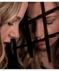 A blonde woman stands in front of a mirror with her gaze lowered. On the mirror, vertical and horizontal lines intersect, drawn with a black marker, resembling barbed wire.
