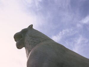 Λήψη από κάτω του μαρμάρινου κεφαλιού μιας λέαινας με ανοιχτό στόμα. Το άγαλμα υψώνεται μεγαλοπρεπές κατω τα αραιά σύννεφα ενός γαλανού ουρανού.