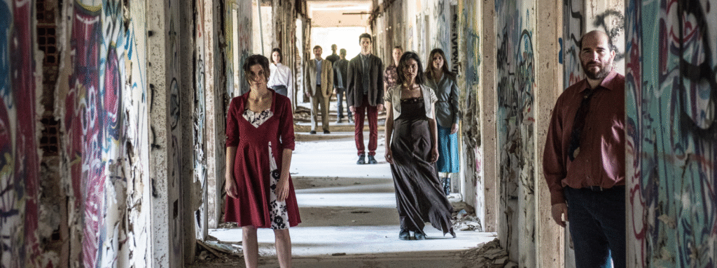 In a long corridor of a derelict building covered in graffiti on the walls and debris in the corners, around 10 people stand scattered across the space, facing us. Their bodies upright and their faces serious as they gaze into the lens.