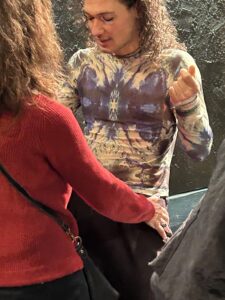 Ένα νεαρό αγόρι με ξανθές μακριές μπούκλες και έντονο μακιγιάζ κινεί τη λεκάνη του μπροστά από μια γυναίκα με κόκκινο πουλόβερ και σπαστά καστανά μαλλιά. Ο νεαρός ντυμένος με διχτυωτό κολλητό μπλουζάκι με ψυχεδελικά μοτίβα της μιλά καθώς εκείνη τον ακουμπά μαλακά στους γοφούς.