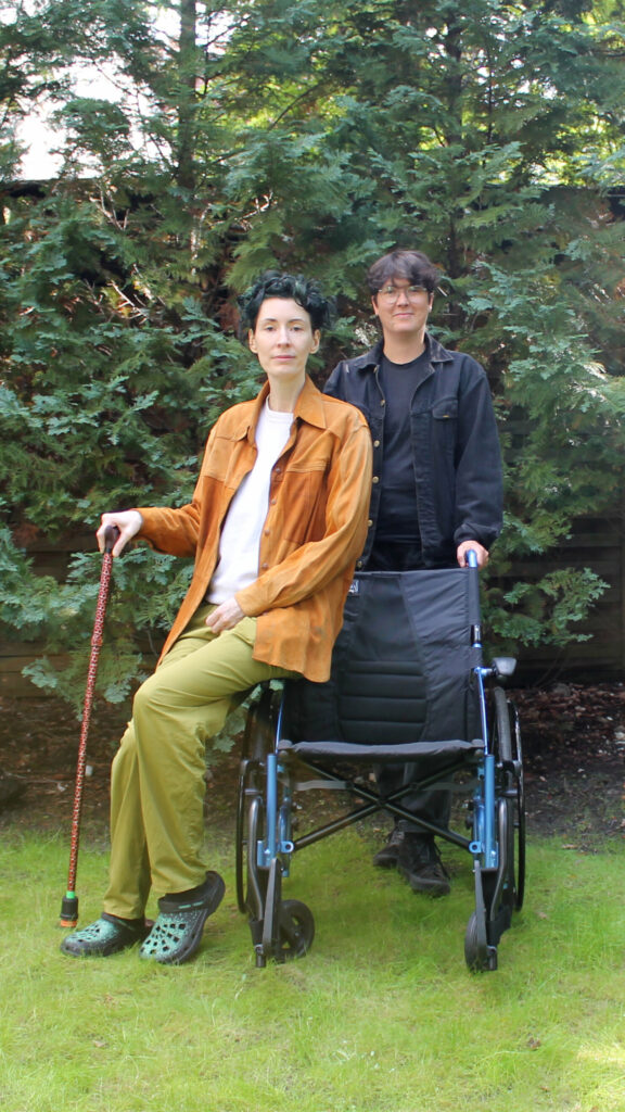 In front of a wooden fence framed by green trees, two people pose with warm smiles. One, dressed in light-colored clothing, has short black hair and rests their right hand on a cane. They appear to be gently seated on the armrest of a wheelchair, holding onto it with both hands. Slightly behind and to the side stands the second person, wearing glasses and black clothing. With short brown hair, they are partially obscured by the wheelchair, their lower body out of view.