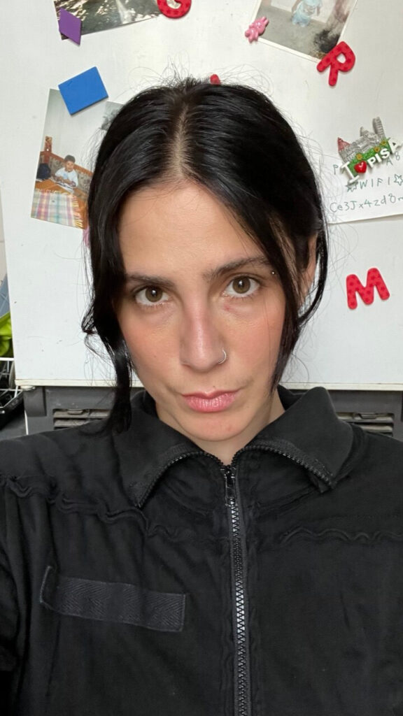 A woman with black hair parted at the waist and tied in a braid sits facing the camera. Her light brown eyes, sharp and unyielding, lock sternly with the lens. A small nose piercing adds a subtle detail to her composed expression. She wears a black sports jacket with a flap, its minimal design contrasting with the domestic setting behind her — faint hints of kitchen objects suggesting she’s seated in a casual, familiar space.