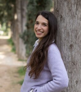 Μία νεαρή κοπέλα με μακριά καστανά μαλλιά ακουμπά την πλάτη της στον κορμό ενός δέντρου σε ένα πάρκο. Το πλατύ της χαμόγελο κάνει τα καστανά της μάτια να φαίνονται μικρότερα. Τα λυτά μαλλιά της πέφτουν από τη μία πλευρά κρύβοντας μέρος του λιλά πουλόβερ της.