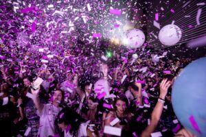 Στιγμιότυπο από το υπαίθριο πάρτυ της Στέγης έξω από το κτίριό της. Ο δρόμος είναι γεμάτος με κόσμο που έχει σχεδόν ολοκληρωτικά καλυφθεί από φούξια και λευκά μακρόστενα κομφετί που έχουν εκτοξευτεί στον ουρανό.