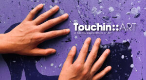 Ένας εικαστικός πίνακας με αφηρημένα σχήματα σε μωβ και μπλε αποχρώσεις παρουσιάζει παχιές πιτσιλιές χρώματος διάσπαρτες στον καμβά. Δύο χέρια αγγίζουν μαλακά την ανάγλυφη επιφάνειά του. Στη δεξιά πάνω πλευρά της φωτογραφίας με λευκά γράμματα είναι γραμμένο: Touching Art: A tactile exploration of Art. Το γράμμα G αποδίδεται με τις τέσσερις τελείες της γραφής braille.