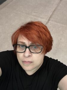 Φωτογραφία πορτρέτου μιας κοκκινομάλλας γυναίκας με γαλάζια μάτια, κοκκάλινα γυαλιά οράσεως και χλωμή επιδερμίδα. Η γυναίκα φορά μαύρη μπλούζα και ποζάρει σοβαρή τραβώντας τη σέλφι από πάνω με τα χέρια της ψηλά.